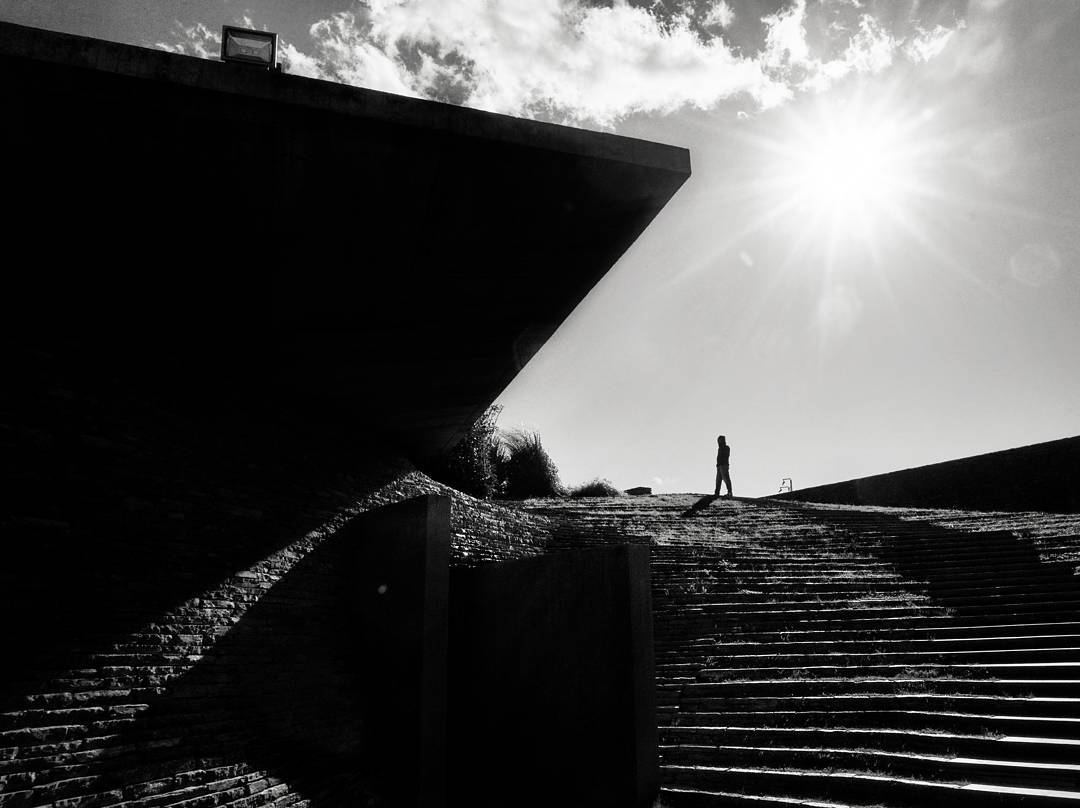 Black-and-white photograph of broad steps, a triangular roof and a lone figure in strong sunlight.